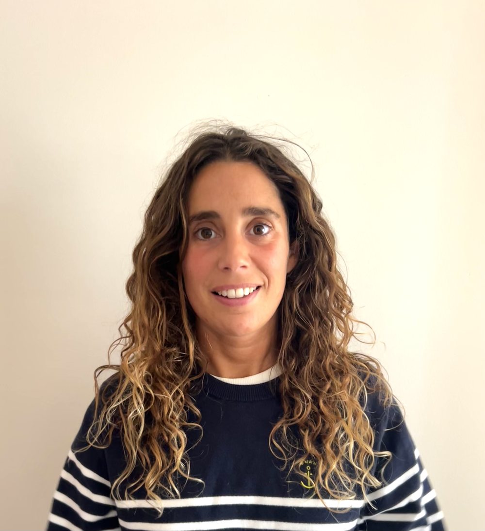 Maria sonriendo, con un jersey azul marino de rayas, delante de una pared crema